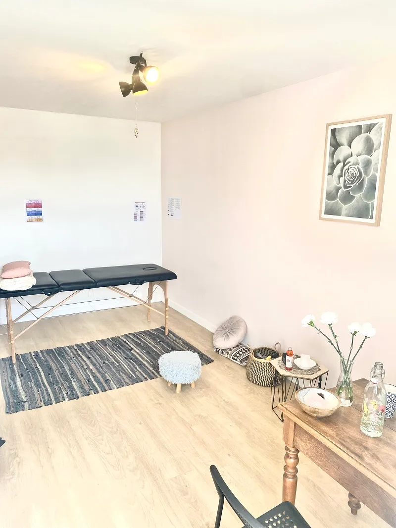 Pièce du cabinet avec table de massage, tabouret, tapis tressé, fleurs blanches sur une table et affiche encadrée d’une plante grasse